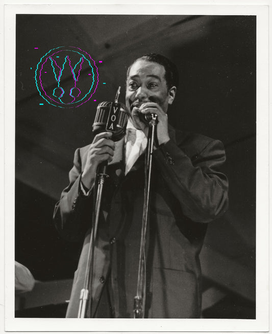 Black and white vintage photograph of a man singing into a microphone on stage, wearing a suit jacket and shirt. The photo has a white border and a colorful circular graphic overlay on the upper left.