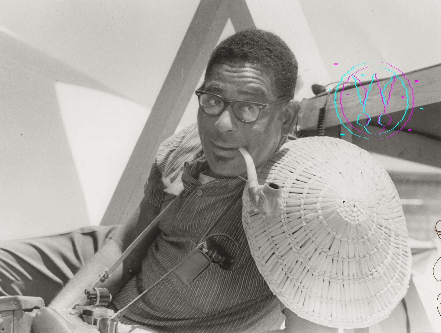 Black and white photograph of a man wearing glasses, a striped shirt, and holding a large woven hat, seated indoors near geometric structures and a microphone.