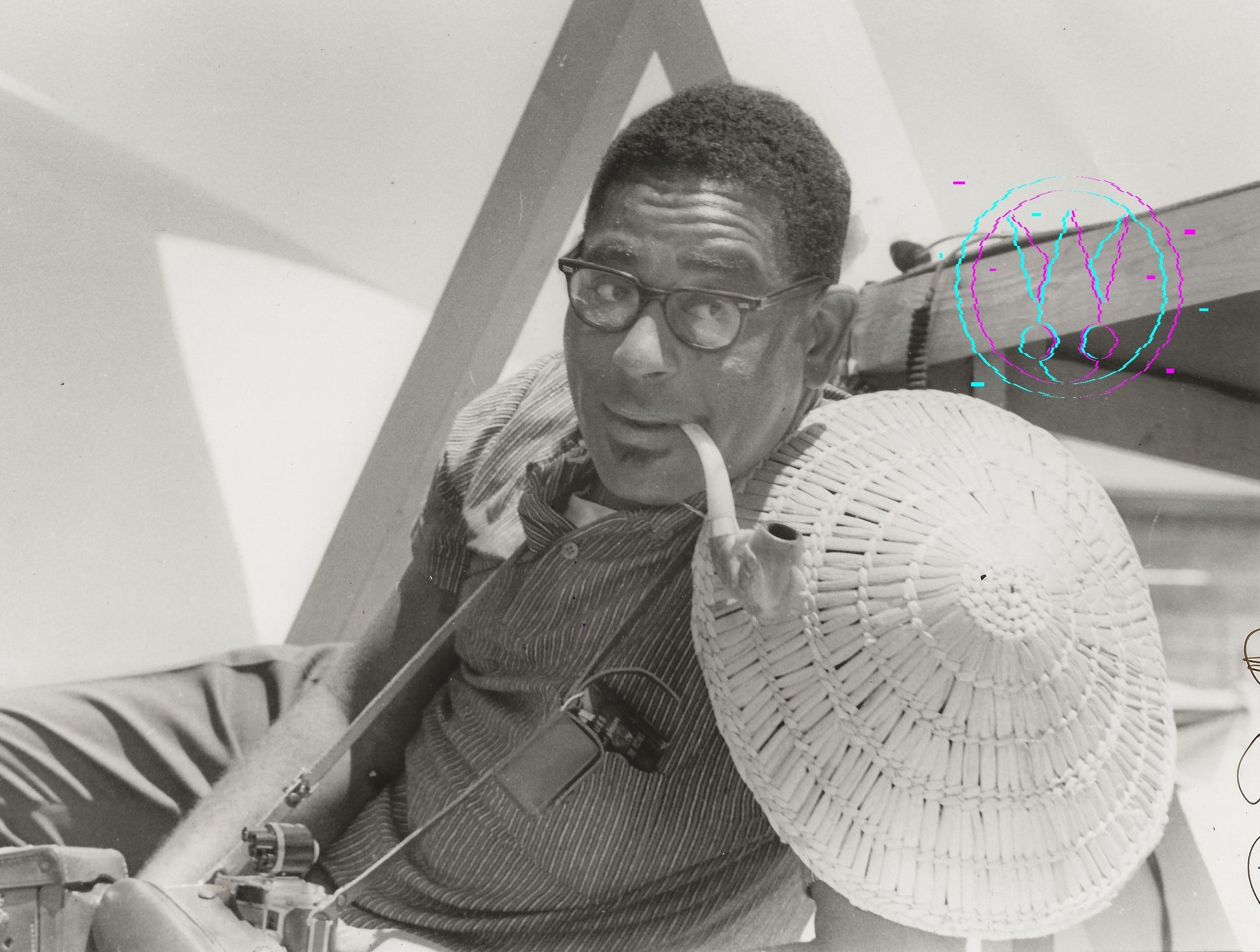 Black and white photograph of a man wearing glasses, a striped shirt, and holding a large woven hat, seated indoors near geometric structures and a microphone.