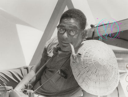 Black and white photograph of a man wearing glasses, a striped shirt, and holding a large woven hat, seated indoors near geometric structures and a microphone.