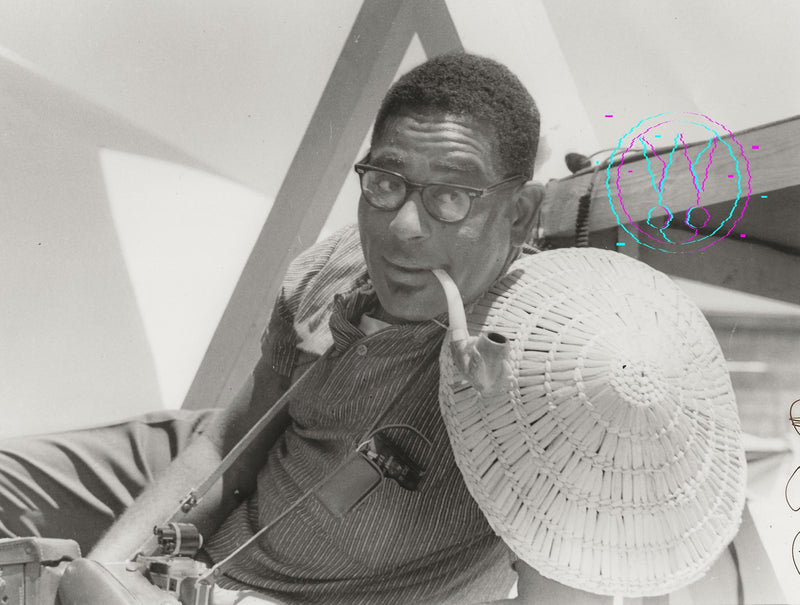 Black and white photograph of a man wearing glasses, a striped shirt, and holding a large woven hat, seated indoors near geometric structures and a microphone.