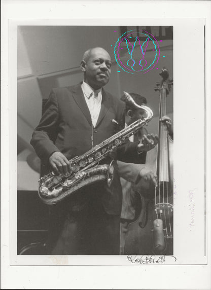 A black and white vintage photograph print of jazz musician Coleman Hawkins playing a tenon saxophone, with a double bass visible in the background. The print features a white border with a handwritten signature at the bottom right and a colorful circular watermark overlay near the top right.